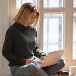 woman in gray sweater sitting on wooden floor typing on portable computer